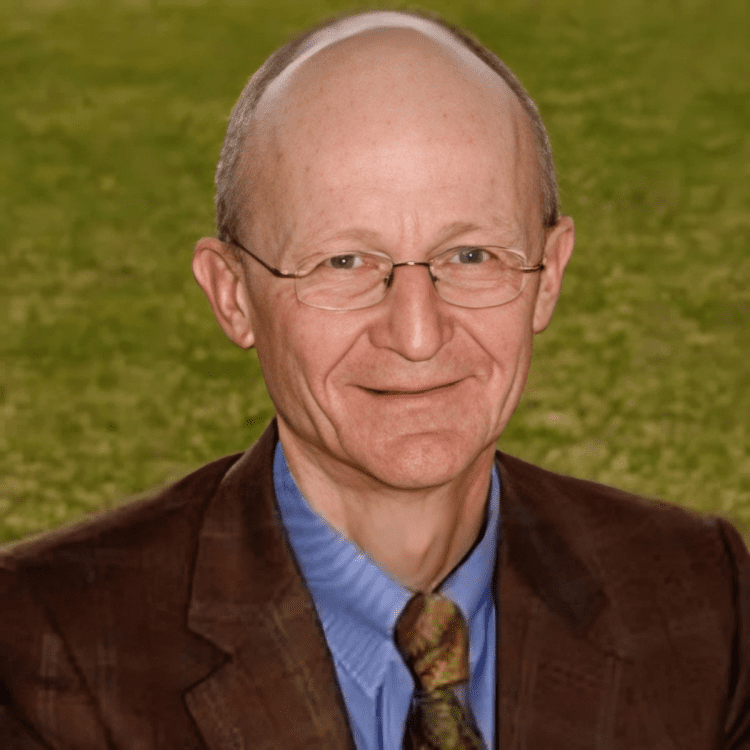 Man in brown suit, glasses, smiling outdoors.